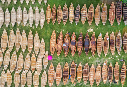 Aerial view of rows of boats resting on green grass create a textured pattern, with a person holding a pink umbrella providing a pop of color, Dhaka, Bangladesh.