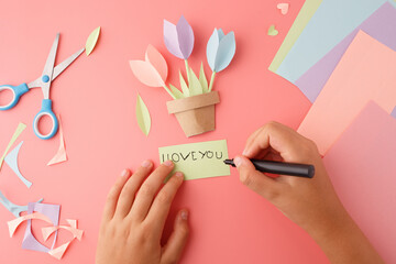 Child creating handmade Mother's Day card with paper tulips and note on pink background, suitable...