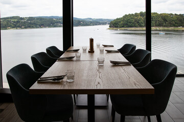 A beautifully set dining table with elegant place settings and glassware, offering a stunning panoramic view of a serene lake and lush green hills through large windows