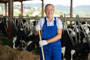 Positive senior man in plaid shirt stands near pole under canopy, weekday rural worker. He is...