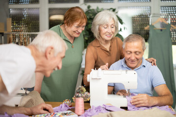 Experienced male sewing teacher sitting at worktable in tailoring workshop, demonstrating basics of...