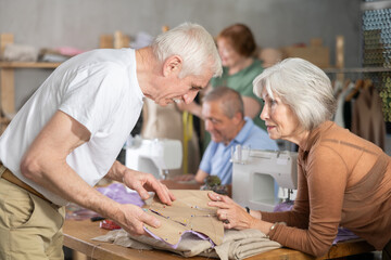 Curious aged woman gesturing to fabric pieces and asking classmate for input, discussing accuracy...
