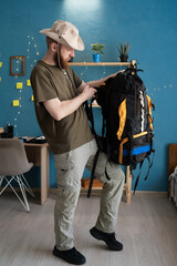 Hiker in hat packing hiking backpack preparing for trip indoors at home. Tourism, travel and hiking