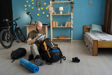 Bearded traveler preparing for hiking trip packing camping gear sitting on floor. Backpack,...