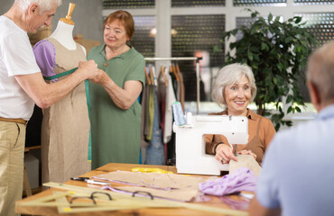 In creative course for seniors, cheerful elderly woman working at sewing machine while other...