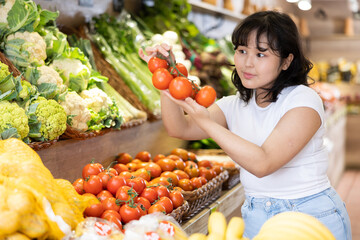 Positive young asian girl selecting ripe red tomatoes while shopping in fruits and vegetables section of supermarket..