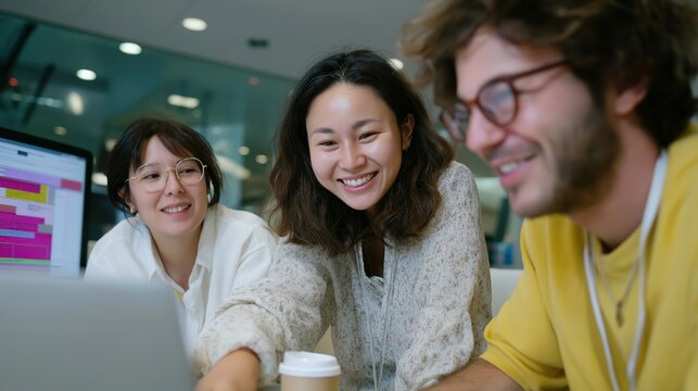 Three people are sitting around a table with a laptop open. One of the women is smiling and pointing at the laptop. The man in the yellow shirt is smiling and looking at the laptop
