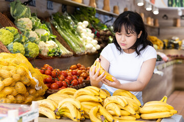 Young girl choosing fresh yellow bananas in fruit and vegetable section of supermarket
