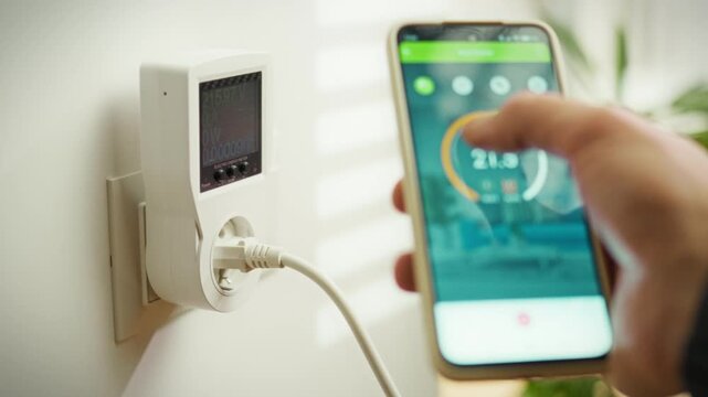 A person is interacting with a smart home energy monitoring device plugged into a wall outlet. The smartphone displays controls and data for managing energy consumption in real-time