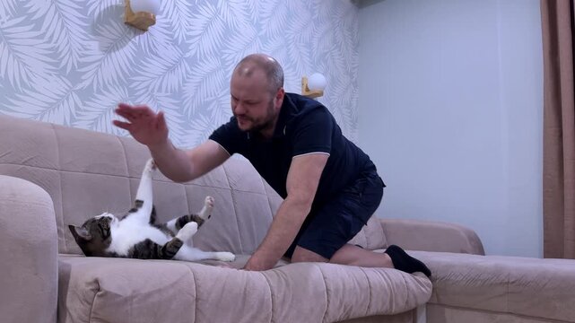 In his room, a man teases his cat with a laser pointer, and the cat chases the red dot with eager focus.