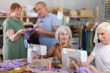Elderly men and women sew on sewing machine and discussing pattern in group during lesson