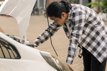 Asian man inspecting the engine under the open hood of a white car, looking concerned and leaning...