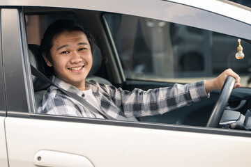 Asian man sitting in the driver's seat of a car with seatbelt on, smiling at the camera, wearing a plaid shirt, holding the steering wheel confidently with a relaxed posture