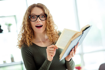 Young woman with glasses enthusiastically reading a book indoors showcasing surprise and excitement...