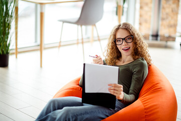 Young woman with glasses working in a modern interior workspace, seated on an orange beanbag and...