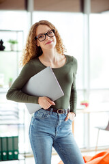 Young woman in casual attire holding a laptop, standing confidently and smiling in a bright office...