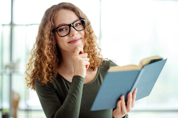 Attractive young woman studying indoors holding a book and smiling thoughtfully in a casual style...