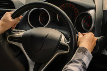 Close-up of an Asian person’s hands holding the steering wheel of a car from the driver’s seat,...