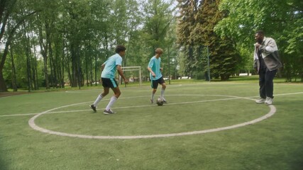 Three young soccer players moving across grassy field with ball as their African American male coach following closely behind, observing their performance during sunny training session in park - Powered by Adobe
