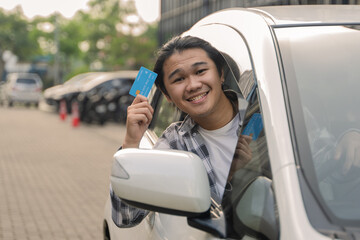 Asian man smiling while showing a blue credit card out of the car window in an outdoor parking area...