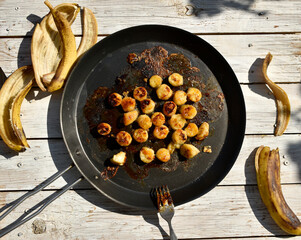 Fried banana slices in caramel on black frying pan on white wooden background. Preparation process