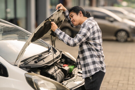 Asian man examining under the hood of a white car with a thoughtful expression in a parking lot, appearing to troubleshoot an engine issue during the day