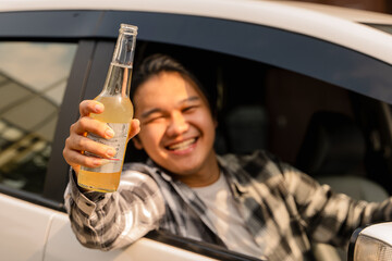 Asian man smiling joyfully from a car window, holding a cold yellow beverage in a glass bottle...