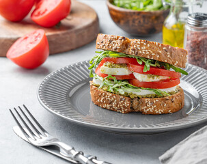 Sandwich of grain bread with fresh tomatoes mozzarella, arugula and pesto on a plate on a light background