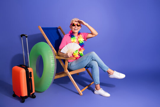 Elderly woman enjoying vibrant vacation setup with summer props, sitting on deck chair against purple background