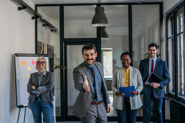 Businessman offering handshake to welcome client in office with team behind him