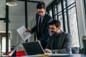 Businessmen working together analyzing financial charts on laptop in office meeting
