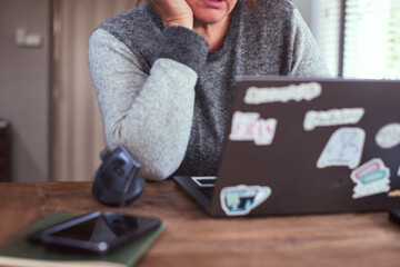 anonymous older woman in a gray sweater resting their chin on their hand at a wooden desk. The desk contains a laptop with stickers, a smartphone, a green notebook, and a computer mouse.