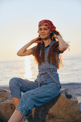 Lifestyle woman in modern boho-western denim street style sitting by the sea wearing red bandana,...