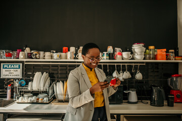 Smiling manager using smartphone and drinking coffee in office kitchen