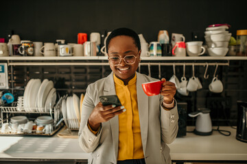 Smiling manager using smartphone and drinking coffee in office kitchen