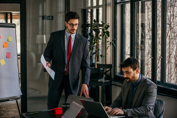 Businessmen discussing charts and working on laptops in modern office