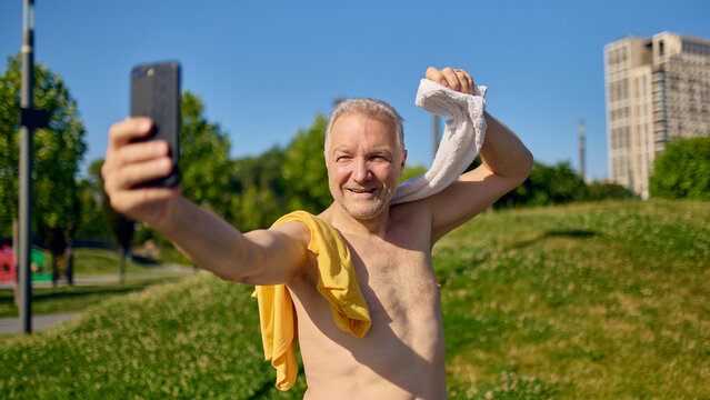 Shirtless senior man smiling and taking selfie with towel after outdoor exercise. Concept of healthy aging, active lifestyle, workout celebration, fitness social sharing, body confidence.