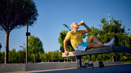 Older man in sportswear drinking water after workout on park bench. Concept of post-training...