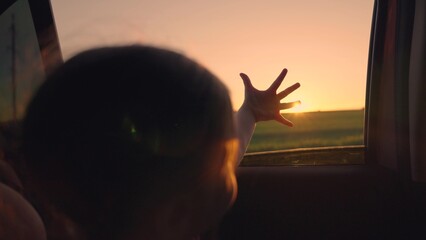 Little daughter dreams, looks out of open car window, child plays with hand sun. Happy family enjoys car ride in fresh air, joint rest. Family car ride. Kid girl sits car, looks out window waves hand