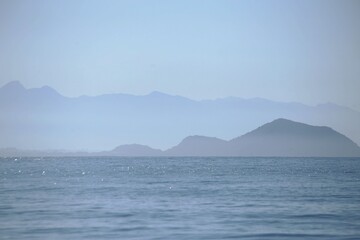 Foggy morning at Prainha Branca in Guaruja, Brazil. blue sea water, blurred horizon line, silhoueete mountains.  Seacape in blue