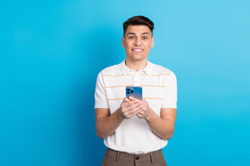 Young man holding smartphone against blue background expressing excitement and wearing casual...