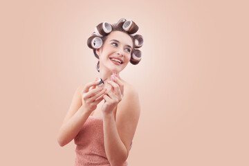 Young smiling woman with hair curlers applying nail polish while looking to the side with her blue...