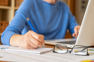 Businesswoman working from home, taking notes and typing on a laptop, with eyeglasses on her nose and financial charts spread across the desk, focusing on analysis and planning