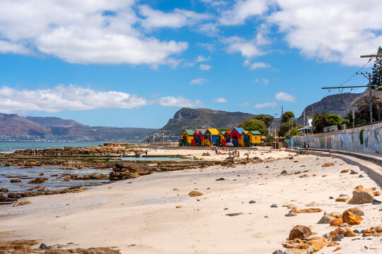 Ein Sandstrand in St. James, Südafrika, mit bunten Umkleidekabinen, einem Meeresschwimmbecken, Felsen und Bergen im Hintergrund unter blauem Himmel