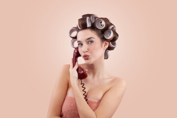 Woman with hair curlers using a vintage telephone while blowing a kiss at the camera. Concept of hairstyle, makeup, beauty routine, and cosmetics. Possibly Making a hair appointment. Isolated on beige