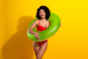 Young cheerful woman in red swimsuit holding green summer ring on yellow background, celebrating sunny vacation vibes