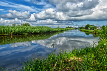 Verdant greenery bordering serene drainage canal, reflecting dynamic cloudscape across typical dutch polder landscape