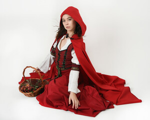 portrait pretty young female brunette model wearing a red hooded fairytale cloak and medieval historical gown. kneeling pose holding wicker basket. Isolated figure, white studio background