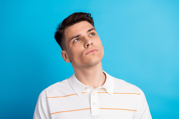 Young thoughtful man in white striped polo shirt looking up against a vibrant blue background...