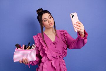 Young woman taking a selfie while holding a makeup bag, dressed stylishly in pink against a vibrant...